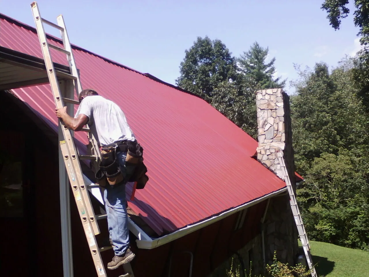 Roofer on a ladder beside a red metal roof and stone chimney — Soffit & Fascia Repair by Roofing Brothers in Middleboro